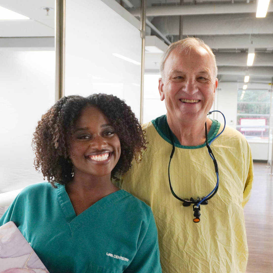 A young female with black curly hair and green scrubs smiling for a photo with a white older man in medical gown standing in a dental clinic
